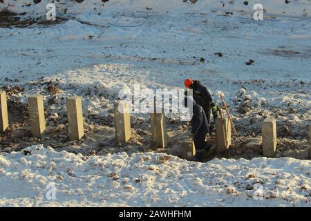 Bauarbeiter in Overalls in orangefarbenen Helmen arbeiten im Winter an einer Baustelle unter Pfählen gegen den Schnee in Russland. Stockfoto