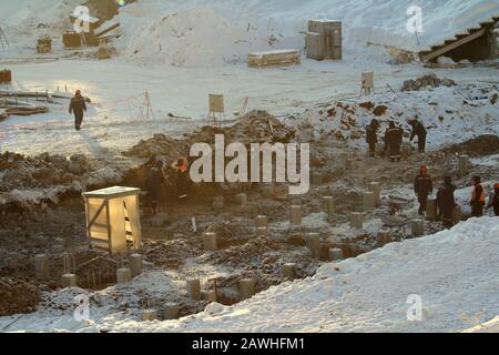 Bauarbeiter in Overalls in orangefarbenen Helmen arbeiten im Winter an einer Baustelle unter Pfählen gegen den Schnee in Russland. Stockfoto