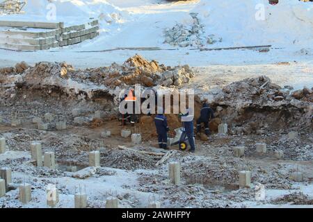 Bauarbeiter in Overalls in orangefarbenen Helmen arbeiten im Winter an einer Baustelle unter Pfählen gegen den Schnee in Russland. Stockfoto