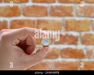Eine Euro-Münze zwischen Daumen und Zeigefinger einer männlichen Hand vor Backstein-Wandhintergrund mit Kopierraum Stockfoto