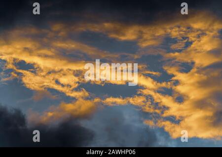 Goldene, flauschige Wolken, die durch verschwindende Strahlen bei Sonnenuntergang und dunkles Kumulonimbus beleuchtet werden, das über sonnigen blauen Himmel schwebt, um das Wetter der Saison zu ändern. Unglaublich Stockfoto