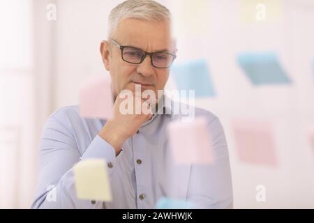 Nachdenklicher leitender Manager, der auf Haftnotizen auf Glas schaut Stockfoto