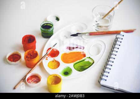 Der Arbeitsplatz des Künstlers - auf einem weißen Tisch sind offene Gläser mit verschiedenen Farben, ein Skizzenbuch, Pinsel, eine Palette, auf der Farben gemischt werden können Stockfoto