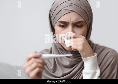 Verunglückte arabische Frau bläst runnige Nase und schaut auf Thermometer Stockfoto