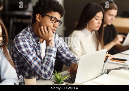 Ermüdete Afro-Kerl, der vor dem Laptop in der Bibliothek sitzt Stockfoto