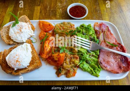 Frühstück mit pochierten Eiern, Toast, Tomaten, Aubergine, Avocado und Speck Stockfoto