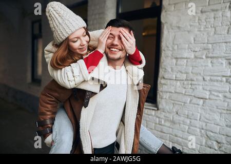 Fröhliche Frau in modischer Mütze und Mantel, die die Augen ihres Mannes mit Handflächen, Glück, Flirt, Zuneigung, Überraschung schließt. Stockfoto