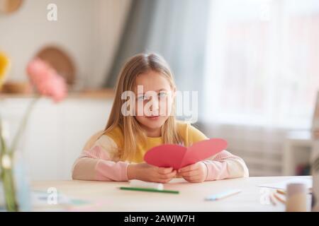 Portrait des niedlichen kleinen Mädchens mit herzförmiger Karte und Blick auf die Kamera, während sie am Tisch im gemütlichen Innenbereich sitzt, Platz für Kopien Stockfoto