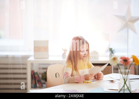 Warmes, gezungtes Porträt eines netten Mädchens, das Urlaubskarte für Muttertag oder Valentinstag macht, während es am Tisch im gemütlichen Innenbereich sitzt, Kopierer Stockfoto