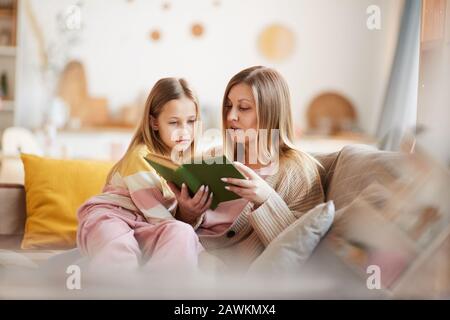Warmtoniges Porträt der reifen Mutter, die Buch zu kleinen Mädchen liest, während sie auf dem Sofa im gemütlichen Innenbereich sitzt, Kopierer Stockfoto
