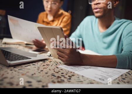 Beschnittenes Porträt zweier ethnischer Geschäftsleute, die spät im dunklen Büro arbeiten, konzentriert sich auf den afrikanischen Mann, der digitales Tablet im Vordergrund hält, den Kopierbereich Stockfoto