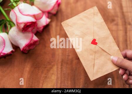 Fröhlicher Valentinstag Hintergrund mit Platz für Kopien. Weibliche Hand, die zum Jubiläum Liebesbrief mit rosafarbenem Rosenstrauß auf einem Holzoberflächenkonzept hält, Stockfoto