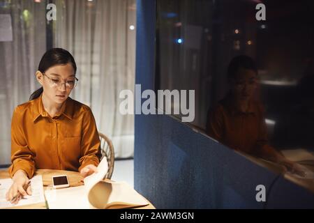 Hochwinkelporträt zeitgenössischer asiatischer Geschäftsfrau, die einen Schreibtisch im dunklen Büroinneren und einen Kopierraum hat Stockfoto