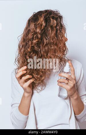 Junge Frau, die ihr lockiges Haar auf weißem Studiohintergrund mit feuchtigkeitsspendenden Sprays besprüht. Stockfoto