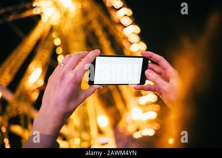 Die Hände der Frau, die das Smartphone mit leerem Bildschirm horizontal halten, fotografieren das Riesenrad im Nachtunterhaltungspark, in der Nähe. Stockfoto