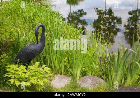 Schwarze Metallreiher Vogelskulptur neben Teich mit Typha Latifolia - Gemeine Cattails und Pondezia cordata - Pickerel Unkraut Im Hinterhof Landgarten Stockfoto