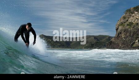 Ein junger männlicher Surfer in einem schwarzen Wetsuit reitet eine Welle vor dem Löwenfelsen am Piha Beach, Piha, West Auckland. Stockfoto