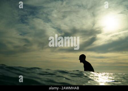 Ein junger männlicher Surfer in einem schwarzen Wetsuit mit einem Ziegenbart sitzt in Silhouette auf einem Surfbrett, das auf eine Welle am Piha Beach, Piha, West Auckland wartet. Stockfoto