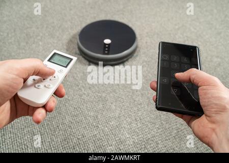 Männliche Hände, die eine Fernbedienung und ein Telefon zur Steuerung eines Roboter-Staubsaugers halten Stockfoto
