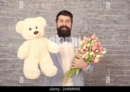 Macho fertig, romantisches Date. Warten auf Darling. Mann gut gepflegt Verschleiß tuxedo Bow Tie halten Blumen Tulpen Bouquet und großen Teddybären Spielzeug. Laden Sie dating. Romantisches Geschenk. Romantischen Mann. Stockfoto