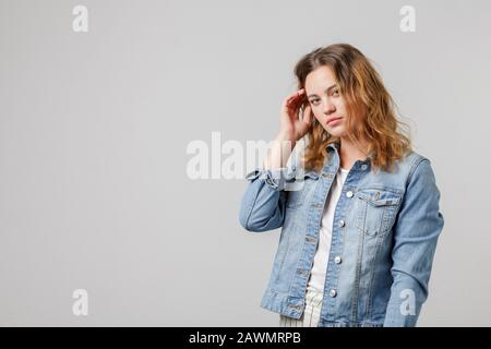 Junges Mädchen mit lang fließenden Haaren in einer Denimjacke sieht nachdenklich an der Kamera aus und berührt ihr Haar. Hochformat in halber Länge auf weißem Hintergrund. Stockfoto