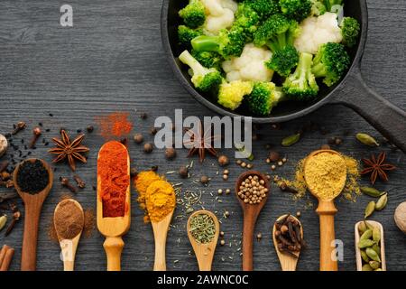 Verschiedene aromatische, bunte Gewürze und Kräuter in Holzlöffeln und Schaufeln. Brokkoli und Blumenkohl auf eiserner Bratpfanne. Draufsicht. Stockfoto