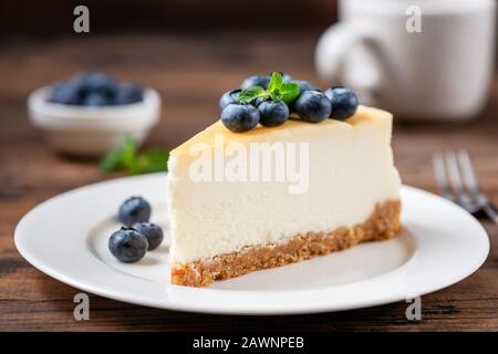 Käsekuchen mit Blaubeeren auf weißem Teller, Holztischhintergrund. Leckeres süßes Dessert. Klassischer New Yorker Cheesecake Stockfoto