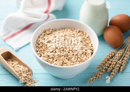 Hafer oder gerollte Haferflocken in Schüssel, Flasche Milch und braune Hühnereier auf blauem Holztisch. Gesundes Frühstück Stockfoto