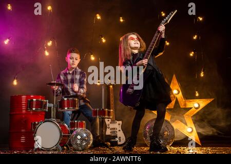 Kinder geben vor, in einer Rockband zu sein und im Studio oder auf der Bühne zu spielen und zu singen. Mädchen spielen an Gitarre und Junge am Schlagzeug. Stockfoto