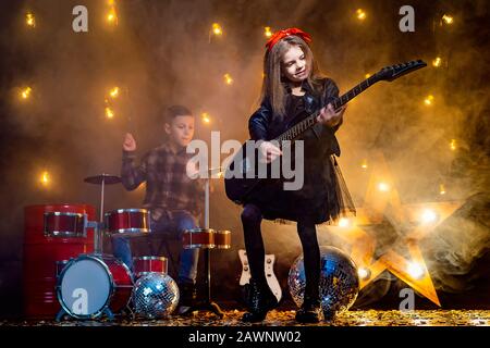 Kinder geben vor, in einer Rockband zu sein und im Studio oder auf der Bühne zu spielen und zu singen. Mädchen spielen an Gitarre und Junge am Schlagzeug. Stockfoto