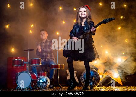 Kinder geben vor, in einer Rockband zu sein und im Studio oder auf der Bühne zu spielen und zu singen. Mädchen spielen an Gitarre und Junge am Schlagzeug. Stockfoto