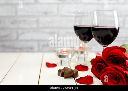 Valentinstag Konzept. Zwei Gläser Wein, rote Rosen, Schokoladenherzen, Rosenblätter und brennende Kerzen auf einem weißen Holztisch mit Platz für Kopien Stockfoto