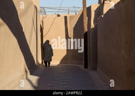 Form einer Frau, die in den Straßen von Yazd, Iran, in der Nähe einer traditionellen Lehmmauer der Altstadt spazieren geht und einen Niqab trägt, ein integrales bescheidenes Gerinnsel Stockfoto