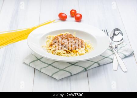 Köstliche Spaghetti-Bolognese mit Fleischsauce mit Tischdecke auf weißem Holz-Plank-Tischplatte mit Rohkostmaterial im Hintergrund. Stockfoto