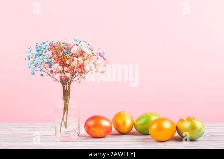 Osterhintergrund mit einem kleinen Blumenstrauß in transparenter Vase und fünf bunten Eiern neben einem rosafarbenen Hintergrund. Stockfoto