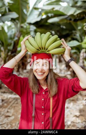 Porträt einer süßen lächelnden Frau, die einen Stamm mit frischen grünen Bananen über dem Kopf hält. Gesundes Ess- und Wellness-Konzept Stockfoto