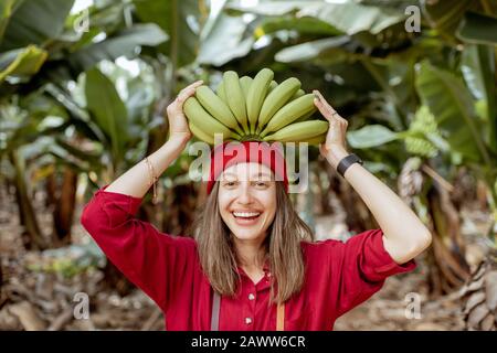 Porträt einer süßen lächelnden Frau, die einen Stamm mit frischen grünen Bananen über dem Kopf hält. Gesundes Ess- und Wellness-Konzept Stockfoto