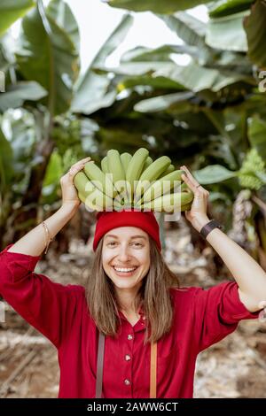 Porträt einer süßen lächelnden Frau, die einen Stamm mit frischen grünen Bananen über dem Kopf hält. Gesundes Ess- und Wellness-Konzept Stockfoto