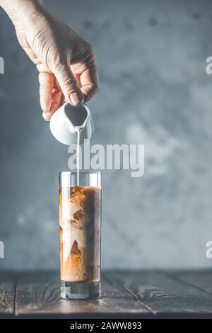 Frauen Hand gießt hausgemachte saure Sahne aus kleinem Glas auf Glas mit kältem Kaffee und Eis. Kaltes Sommergetränk auf dunklem Holztisch und grauem Bac Stockfoto
