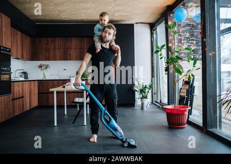 Der junge Vater saugt den Boden der Wohnung mit seinem Baby auf dem Hals Stockfoto