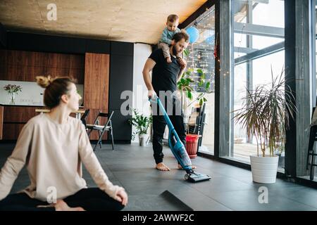 Tägliche Familienaktivität, Kind am Hals des Vaters. Er saugt, sie macht Yoga. Stockfoto