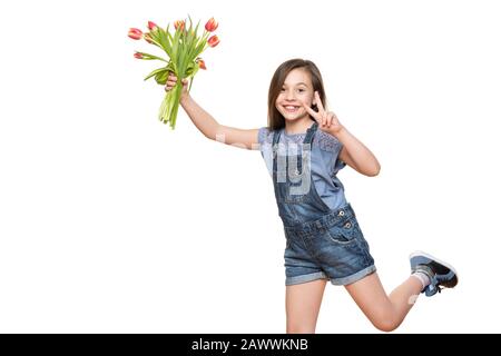 Liebenswürdige Schulmädchen, die mit Blumenstrauß aus Tulpen auf weißem Hintergrund läuft, Kamera betrachtet und das Friedenshandzeichen macht. Stockfoto