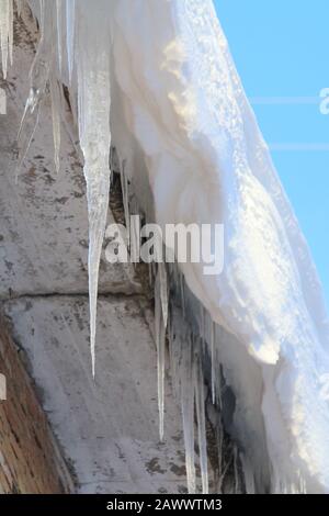 Große Eiszapfen, die vom Dach des Ziegelhauses auf blauem Himmel hängen. Untere Ansicht. Stockfoto mit leerem Textbereich. Stockfoto