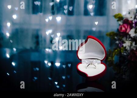 Rote, herzförmige Samtbox mit eingehöhltem Rautenring im Hintergrund mit Bokeh-Lichtern und Blumen. Valentinshintergrund. Geschenke für Frau am Valentinstag. Liebe, Angebotskonzept. Stockfoto