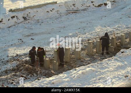 Bauarbeiter in Overalls in orangefarbenen Helmen arbeiten im Winter an einer Baustelle unter Pfählen gegen den Schnee in Russland. Stockfoto