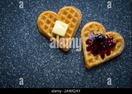 Einige herzförmige Waffeln mit Beerenmarmelade und Butter auf grauem Steingrund Stockfoto