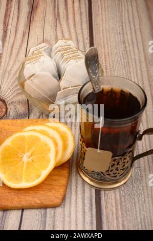 Tee aus facettiertem Glas in einem Vintage-Cup-Halter, Teebeutel in einer Glasvase und geschnittene Zitrone auf einem Brett auf Holzhintergrund. Nahaufnahme Stockfoto