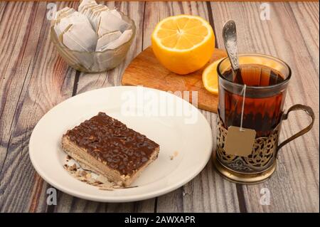 Tee aus facettiertem Glas in einem Vintage-Cup-Halter, Teebeutel in einer Glasvase, geschnittene Zitrone auf einem Brett und ein Stück Waffelkuchen auf einem weißen Teller auf einem Woo Stockfoto