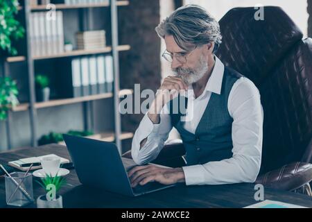 Nahporträt seines hübschen, attraktiven, gutaussehenden, grauhaarigen Gentleman, der im Stuhl sitzt und Daten auf dem Laptop im industriellen Loft-Stil sucht Stockfoto