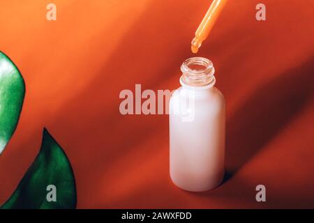 Ölserum mit einem Tropfen, der auf orangefarbenem Hintergrund mit grünem Monstera-Blatt in die Flasche fällt. Spa Kosmetik Hautpflege Konzept Stockfoto